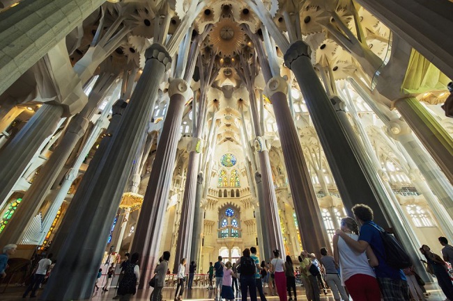 Emberek sétálnak a Sagrada Família belsejében. Magas oszlopok és színes üvegablakok lenyűgöző látványt nyújtanak.