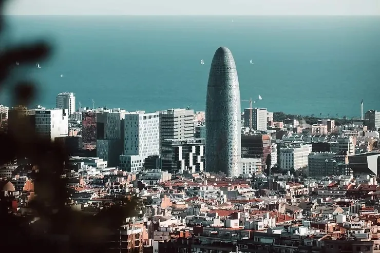 Barcelona városképe, középpontban a híres Torre Glòries toronyház. Háttérben kék tenger, vitorlás hajókkal. Városi élet nyugodt hangulata.