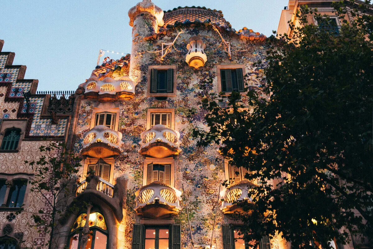 Gaudí festői épülete, a Casa Batlló Barcelonában, este megvilágítva, díszes homlokzattal és erkélyekkel. Színes mozaikok borítják, mellette fák, hangulatos látvány.