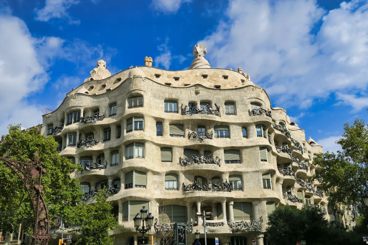 Hullámos homlokzatú épület világos kőből, kovácsoltvas erkélyekkel. Gaudí Pedrera háza Barcelonában. Kék ég háttérben, napfényes, modernista stílus.