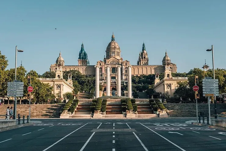 A kép egy impozáns épületet ábrázol Barcelonában, a Katalán Nemzeti Művészeti Múzeumot, parkokkal és szökőkúttal körülvéve, tiszta ég alatt. Elegáns és nyugodt hangulat.