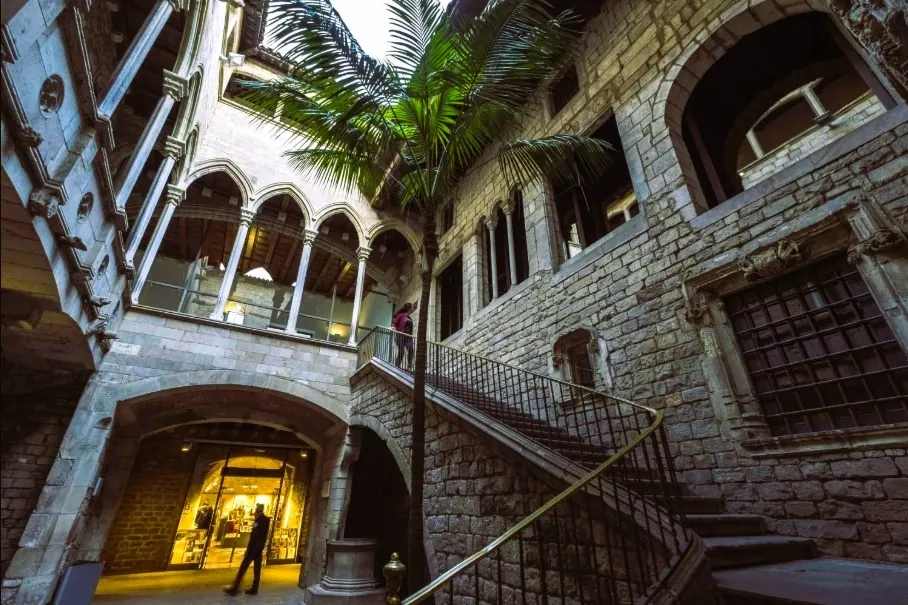Két ember a barcelónai Picaso múzeum történelmi épületének kőudvarában, íves ablakokkal. Egyikük felmegy a lépcsőn. Bent meleg fények, középen pálmafa.