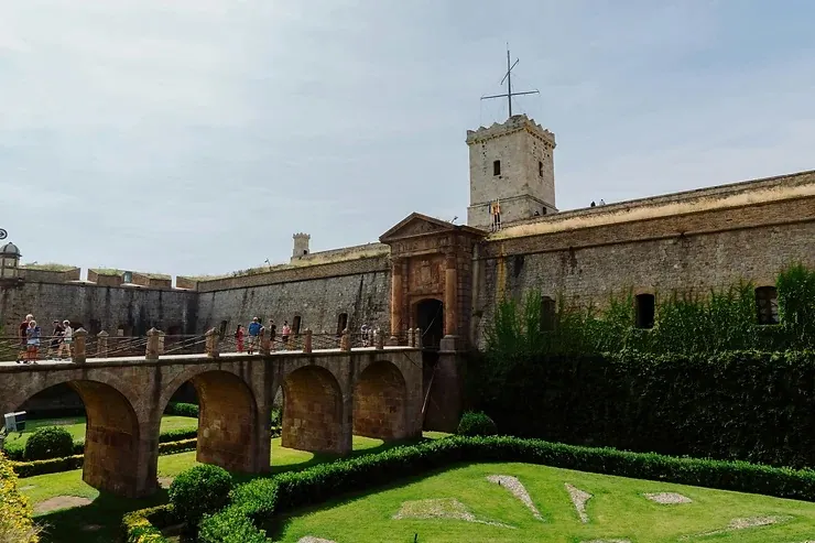 Kőhíd a barcelonai Montjuïc várához vezet. Látogatók sétálnak a hídon. Körülötte zöld kert, a háttérben az ég világos.