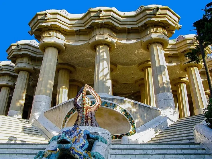 Park Güell kőoszlopai és színes mozaikos gyík szobor napfényben. Széles lépcsők vezetnek felfelé, kék ég háttérrel.