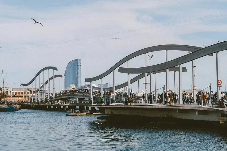 Rambla de Mar híd Barcelonában emberekkel, háttérben a W Barcelona hotel. Kék ég, madarak repülnek, nyugodt, tengerparti hangulat.