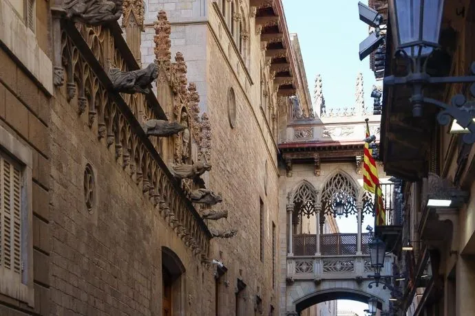Szűk utcán faragott kő épületek, középkori stílusú csúcsíves híd köti össze őket Barcelona Gótikus negyedében. Jobbról piros-sárga zászló lóg, nyugodt légkör.