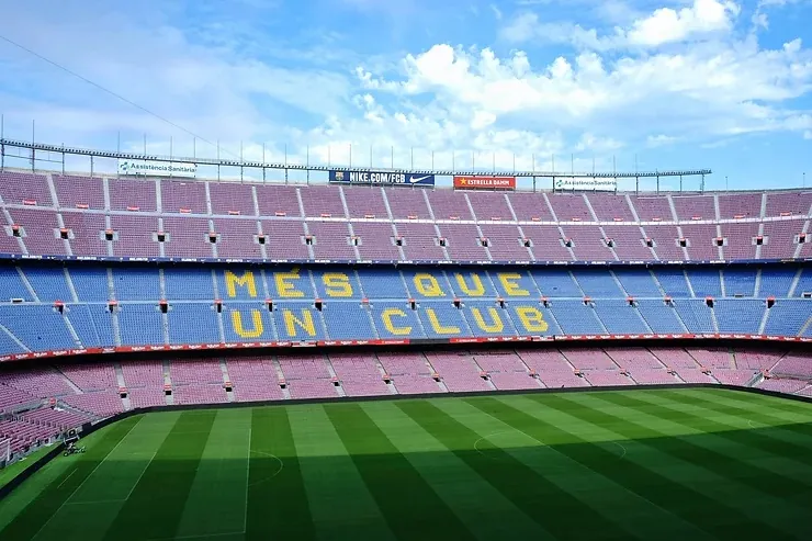 Üres Camp Nou stadion belső tere kék és piros széksorokkal. Középütt sárga felirat: MES QUE UN CLUB. Zöld gyep és kék ég látható.