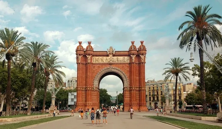 Vörös téglás diadalív Barcelonában, emberek sétálnak alatta. Pálmafák keretezik, kék ég a háttérben. Életvidám hangulat.