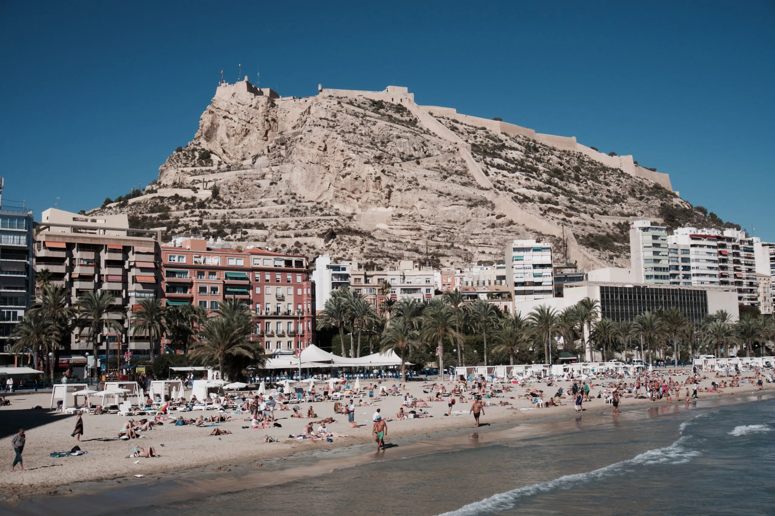 Alicante tengerpart, Playa Postiguet strand napozókkal, háttérben a domb és Santa Barbara vár
