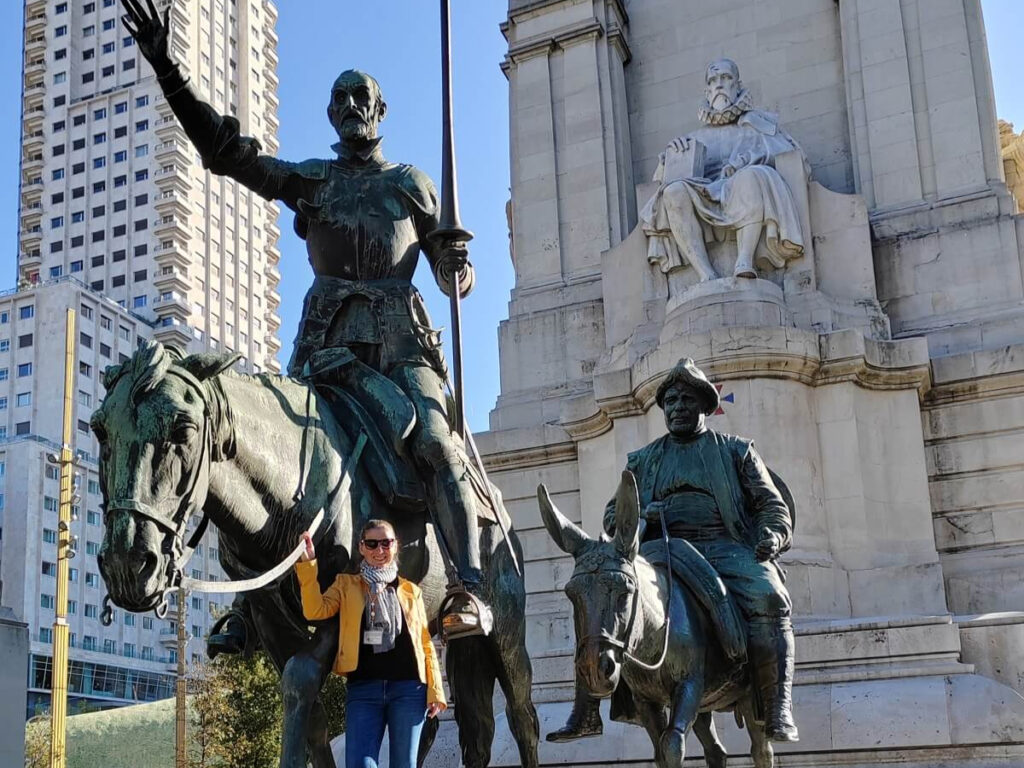 Cervantes emlékmű Madridban, 3 szoborral: lovas alak - Don Quijote, mellette szamárháton Sancho Panza. Hátul szobor, a férfi Cervantest ábrázolja, és modern épület van a háttérben. Nő, Kása Heni madridi magyar idegenvezető sárga kabátban pózolva mosolyog.
