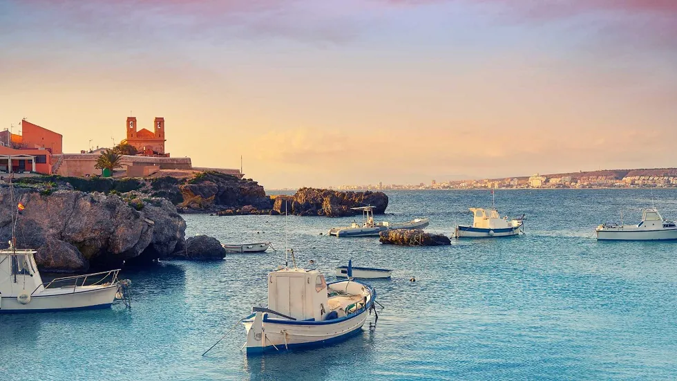 Alicante hajókirándulás Tabarca szigetére. Hajók horgonyoznak a kék tengeren egy sziklás part közelében, háttérben egy vörös épület. Naplemente meleg fényével vonja be a tájat.