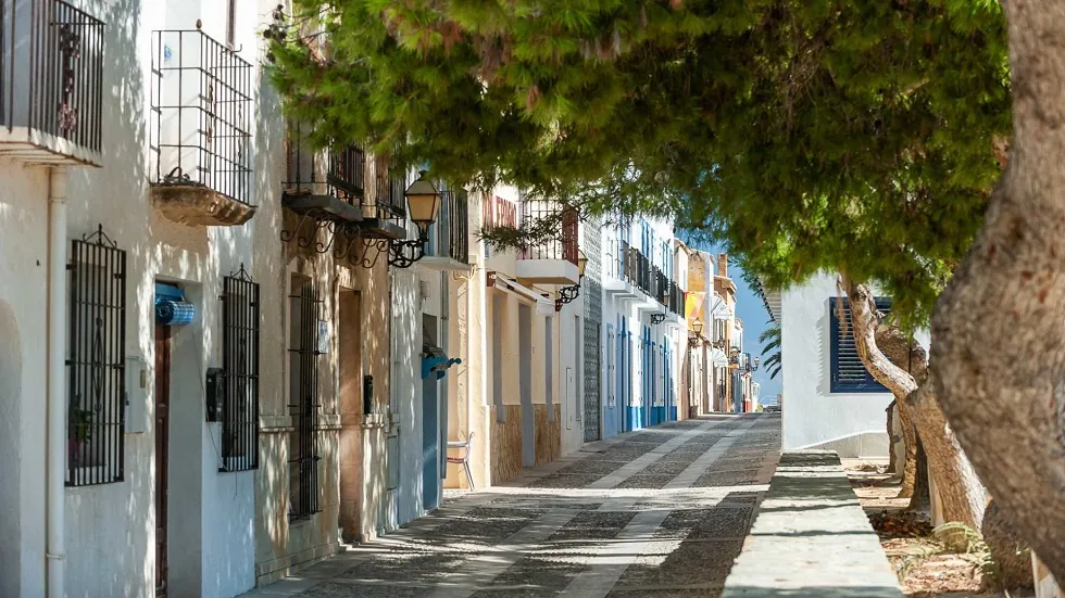 Szűk mediterrán utca Tabarca szigete, fehér és színes házakkal, kék ajtókkal, dús lombú fákkal árnyékolva. Nyugodt, napsütéses hangulat.