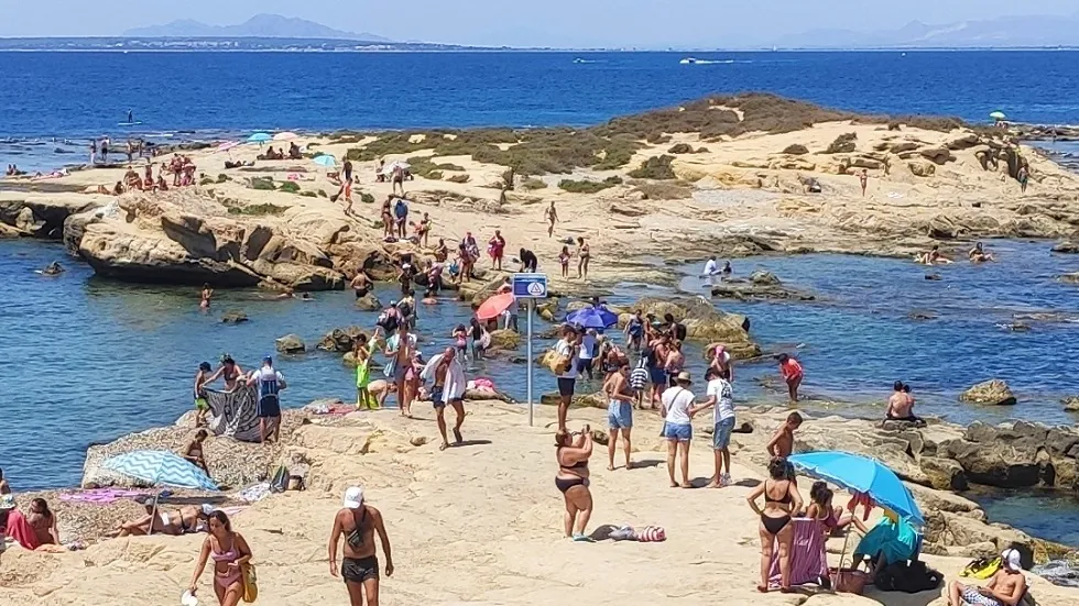Tabarca szigete, tengerparti jelenet kavicsos parton sok fürdőzővel, türkizkék víz, színes napernyők, távolban homokdűnék és hegyek.